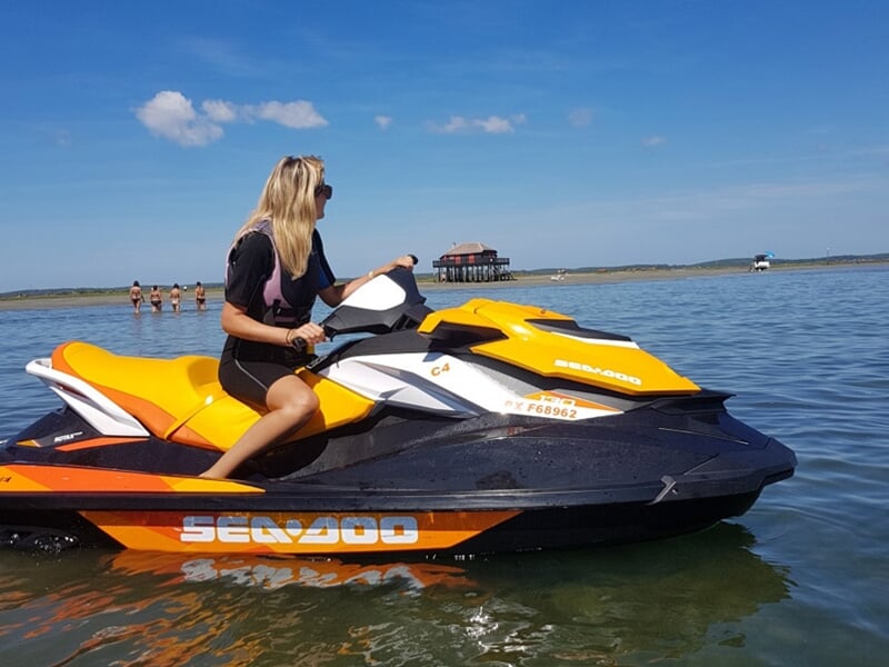 Une femme blonde en gilet de sauvetage sur un jetski jaune et noir dans une eau calme avec une cabane sur pilotis au loin.
