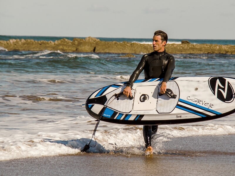 Homme en combinaison noire tenant une planche de surf électrique décorée de bleu et blanc, marchant sur le sable mouillé.
