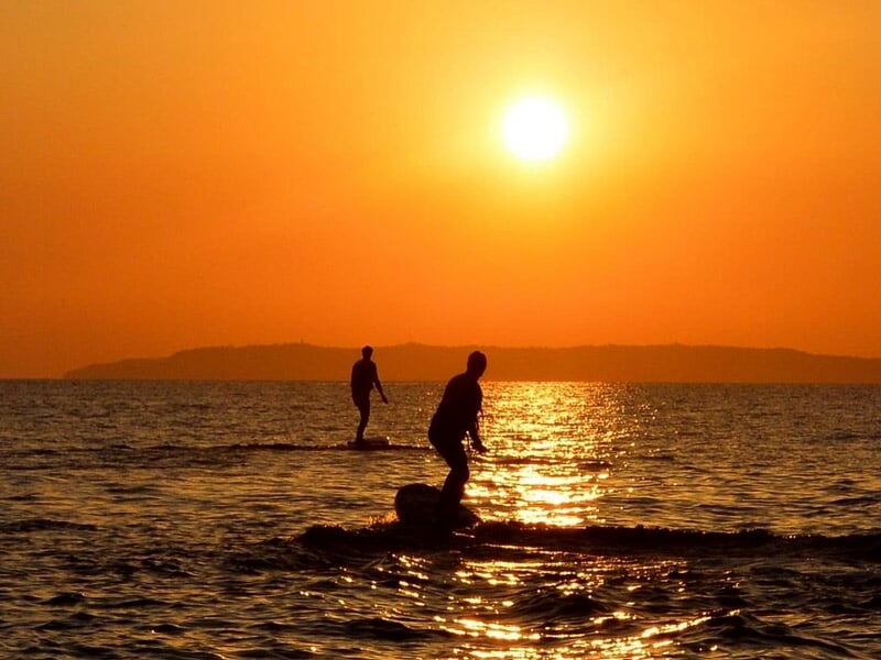 Deux silhouettes sur des planches de foil glissent sur la mer au coucher du soleil avec reflets dorés sur l’eau.