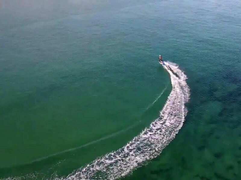 Vue aérienne d’une planche électrique traçant un long sillage blanc sur la mer verte et calme.