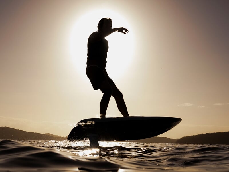 Surfeur debout sur une planche de foil électrique au-dessus de l’eau, silhouette sombre devant un soleil éclatant bas sur l’horizon.