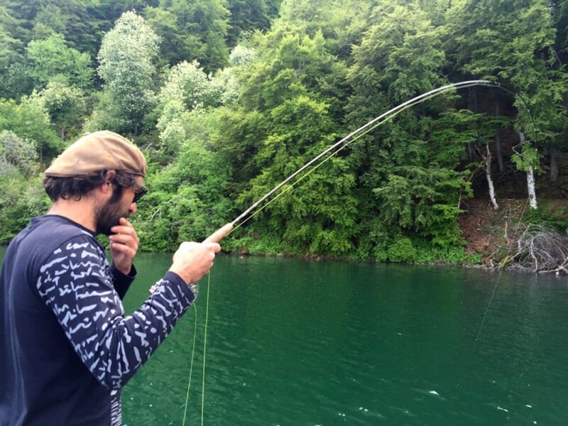 Homme en casquette pêchant à la ligne sur un lac entouré d'arbres verts et d'une forêt dense.