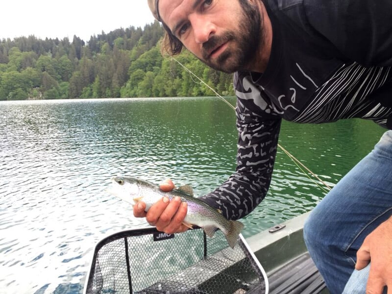 Homme barbu montrant une truite pêchée au bord d'un lac avec forêt en arrière-plan.