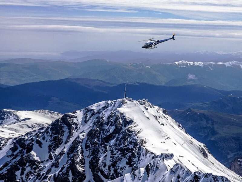 Hélicoptère survolant un sommet enneigé avec des montagnes et vallées en arrière-plan sous un ciel nuageux.