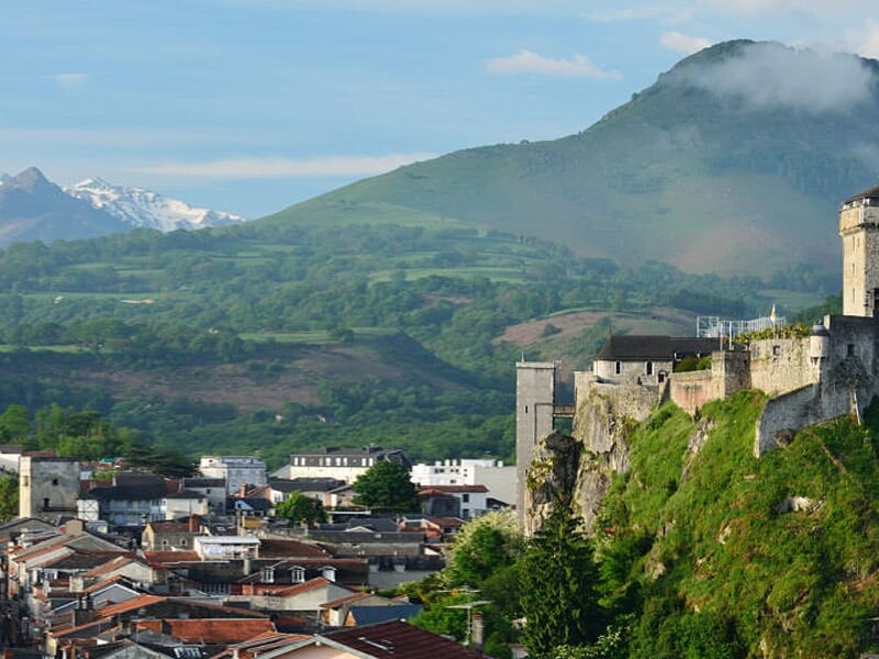Vue panoramique d'une ville avec un château fort sur une colline verdoyante et des montagnes en arrière-plan.