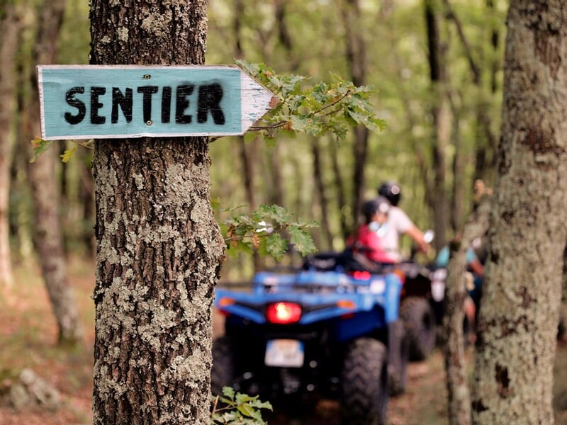 Panneau en bois indiquant un sentier dans une forêt avec un groupe de personnes en quad en arrière-plan.