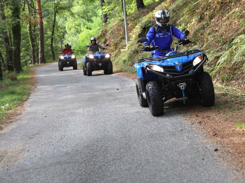 Trois personnes en quad bleu roulant sur une route goudronnée bordée d'arbres et de végétation.