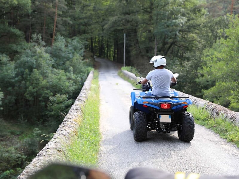 Personne en casque blanc conduisant un quad bleu sur un chemin étroit avec des arbres et un petit pont en pierre.