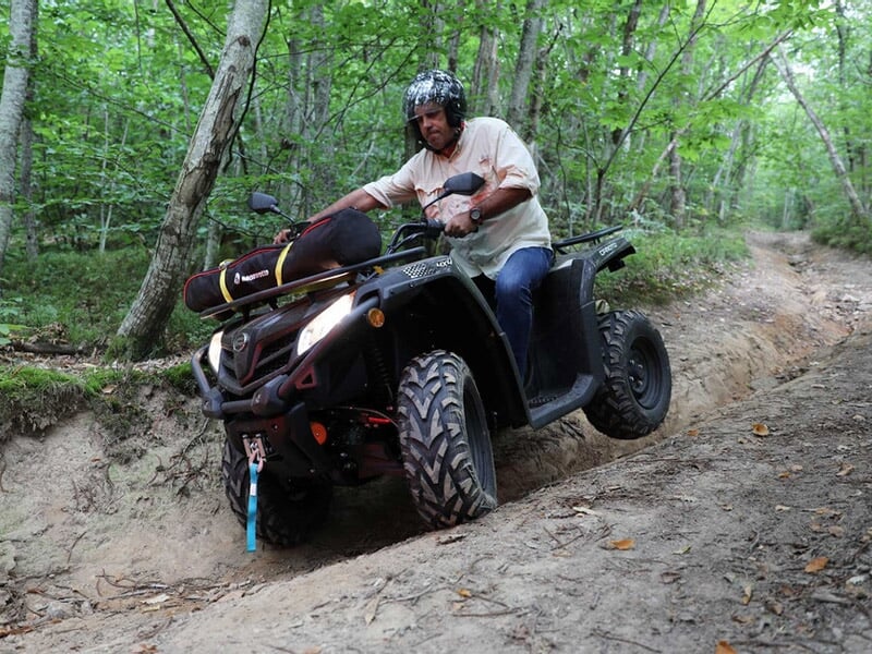 Homme conduisant un quad noir sur un chemin boueux en forêt avec un sac attaché à l'avant du véhicule.