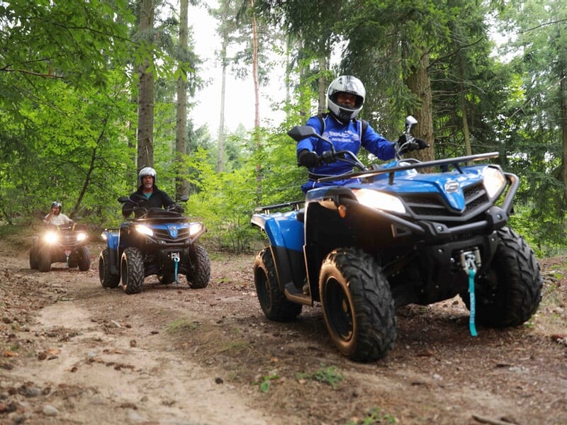 Groupe de personnes en quad bleu roulant sur un sentier forestier avec des phares allumés et des arbres verts autour.