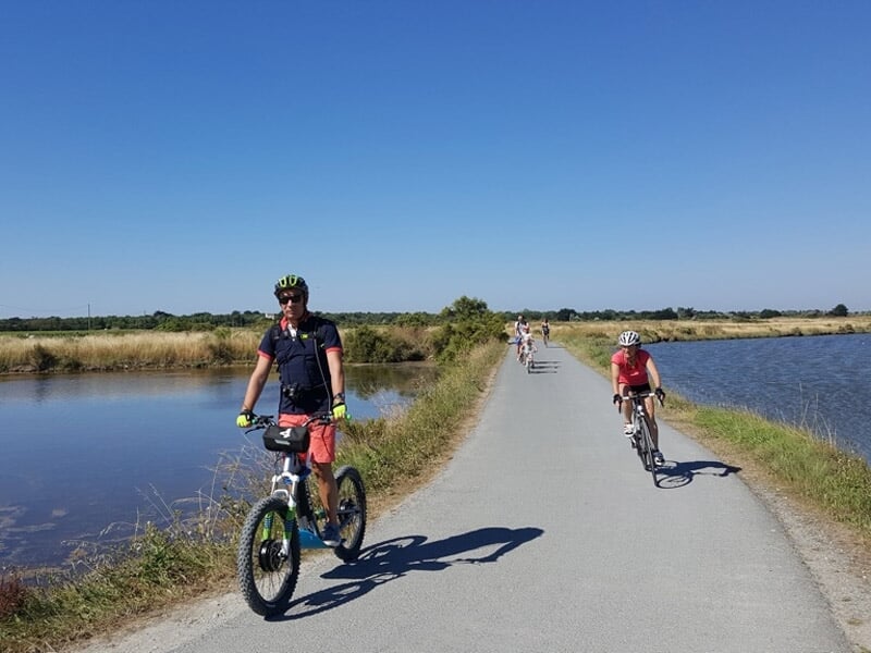 Personne en tenue de sport faisant du vélo électrique sur un chemin entre deux plans d'eau sous un ciel bleu clair.