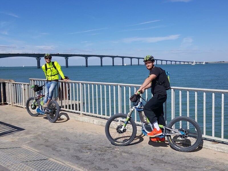 Deux cyclistes posant avec leurs vélos électriques devant une rambarde, avec un grand pont et la mer en arrière-plan.