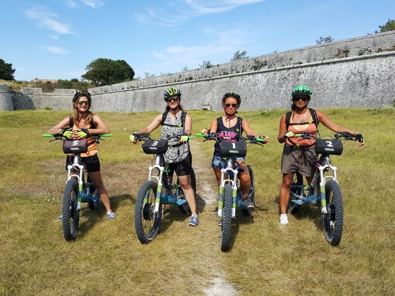Quatre femmes souriantes posant avec leurs vélos électriques sur une pelouse devant un mur en pierre sous un ciel bleu.