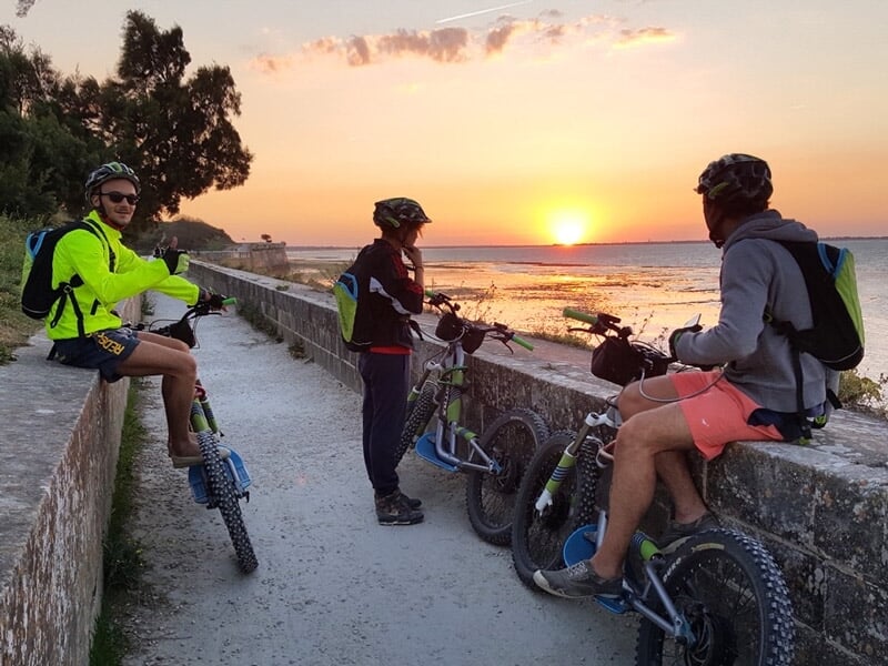 Trois cyclistes en tenue décontractée avec leurs vélos électriques regardant un coucher de soleil sur la mer depuis un chemin côtier.