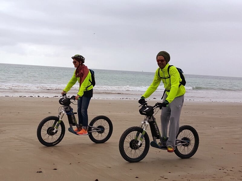 Deux hommes en vêtements de sport roulant à vélo électrique sur une plage de sable avec la mer en arrière-plan sous un ciel nuageux.
