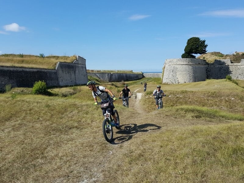 Quatre cyclistes en tenue sportive roulant à vélo électrique sur un chemin herbeux près d'anciennes fortifications en pierre.
