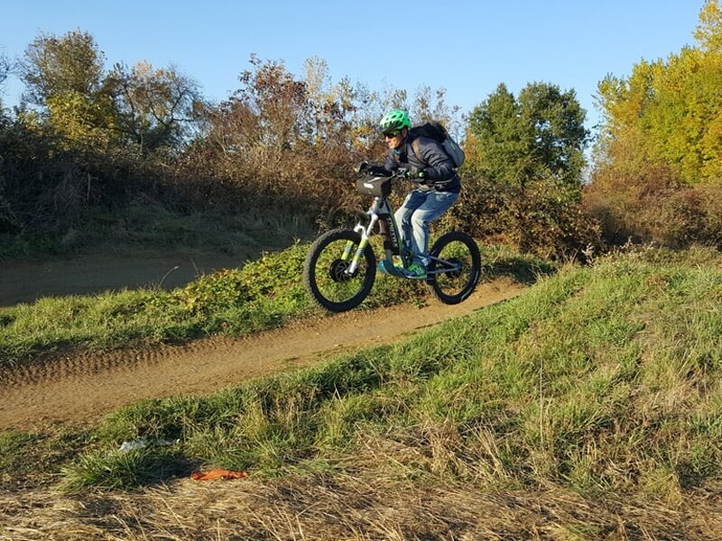 Cycliste en tenue décontractée sautant avec son vélo électrique sur une piste en terre entourée d'herbe et d'arbres verts.