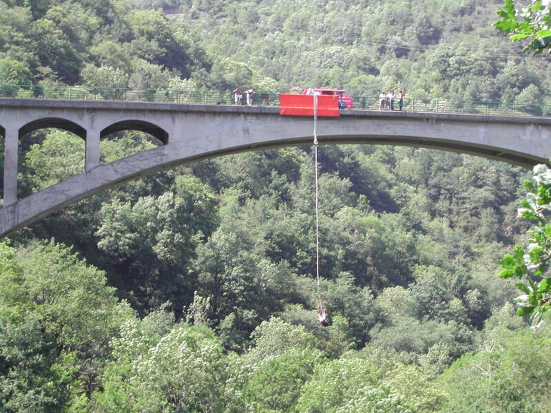 Vue d'ensemble d'un saut à l'élastique depuis un pont en arc, avec un camion rouge et des spectateurs observant depuis le sommet, au-dessus d'une vallée boisée.