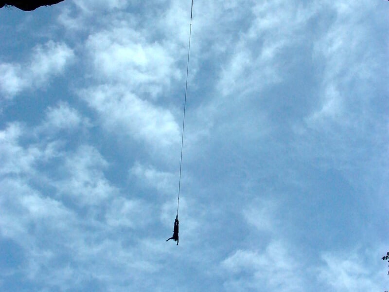Personne suspendue dans le vide par un élastique, en plein saut sous un ciel bleu légèrement nuageux, mettant en avant l'activité extrême du saut à l'élastique.