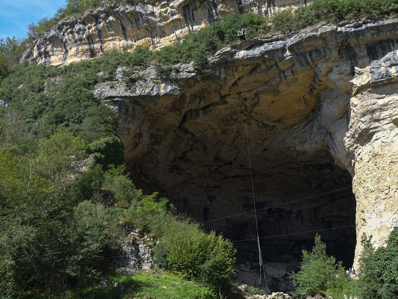 Grotte imposante entourée d'arbres et de rochers, avec un ciel partiellement nuageux.