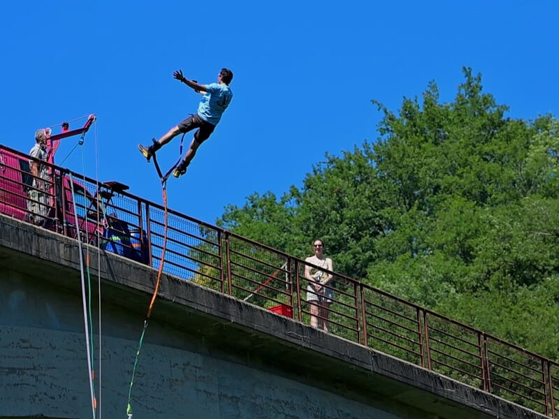 Un homme en train de sauter à l'élastique depuis un pont avec un ciel bleu et des arbres verts en arrière-plan.