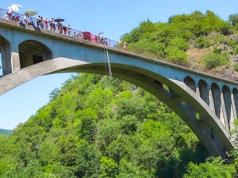 Vue d'un pont en arc avec des personnes sur le dessus et une corde de saut à l'élastique suspendue au-dessus d'une vallée boisée.