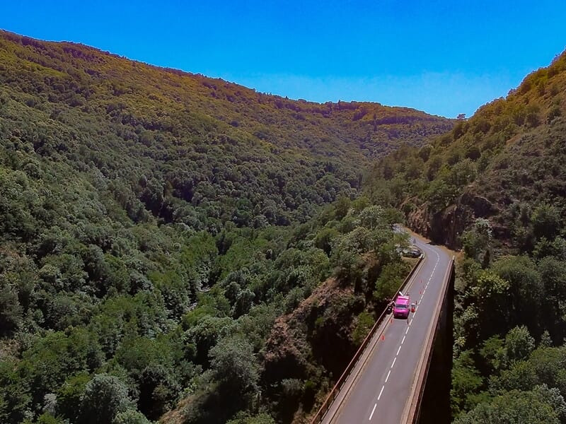 Route sinueuse traversant une vallée dense en forêt avec une voiture rose roulant sur la route sous un ciel clair.