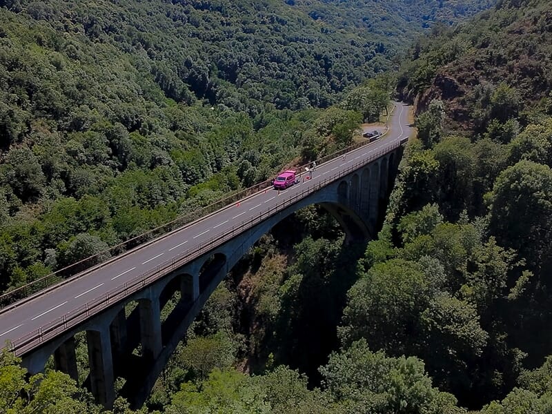 Voiture rose traversant un pont en arc au-dessus d'une vallée boisée avec une route qui serpente dans un paysage naturel.