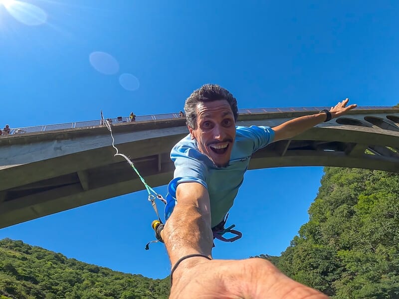 Homme souriant en train de faire un saut à l'élastique, tenant une caméra selfie avec un pont et un ciel bleu en arrière-plan.