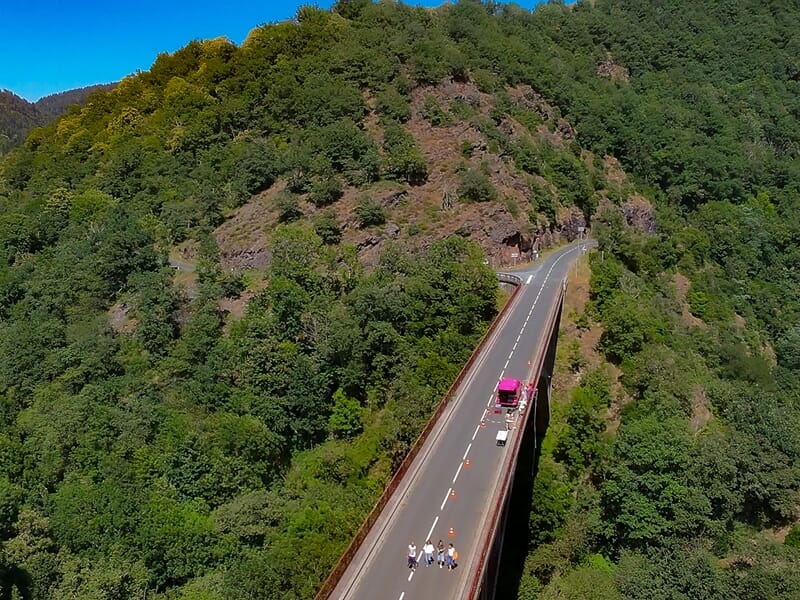 Vue aérienne d'une route traversant une vallée boisée avec une voiture rose et plusieurs personnes sur la route sous un ciel bleu.