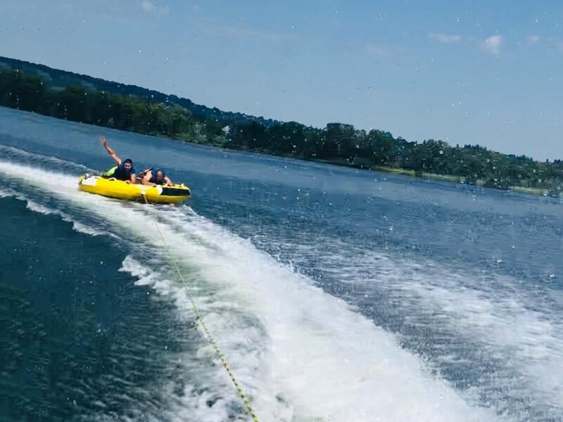 Deux personnes s'amusent sur une bouée tractée jaune sur un lac, éclaboussures d'eau autour.
