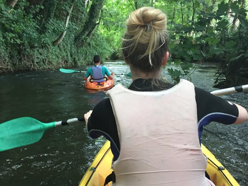 Vue arrière d'une personne pagayant en kayak sur une rivière étroite bordée d'arbres.