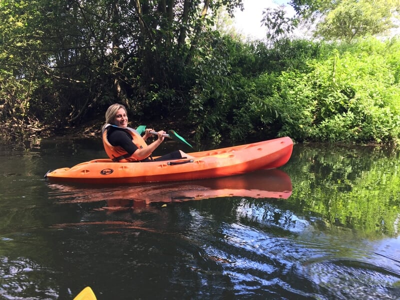 Femme souriante pagayant seule en kayak orange sur une rivière calme et verdoyante.