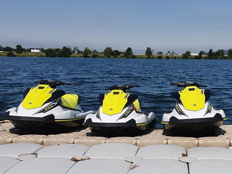 Trois jetskis jaunes alignés sur un ponton flottant au bord d'un lac sous un ciel clair.