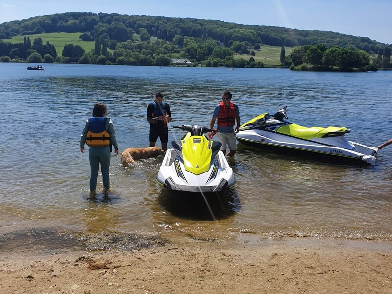 Trois personnes debout au bord d'un lac avec deux jetskis jaunes sur la plage sableuse.