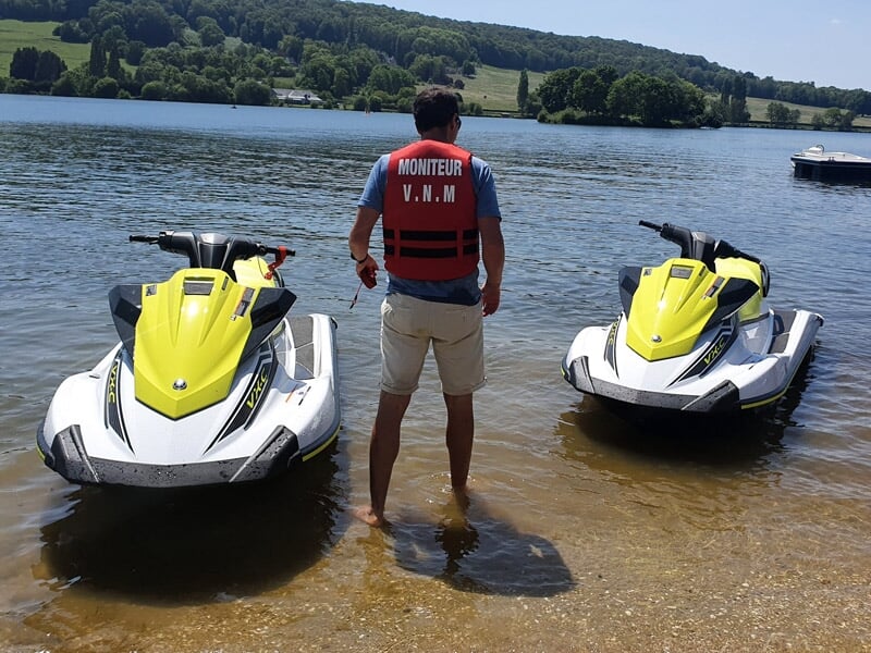 Moniteur en gilet rouge debout entre deux jetskis jaunes sur une plage au bord d'un lac.