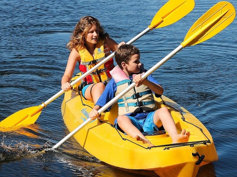 Deux enfants en gilets de sauvetage pagayant dans un kayak jaune sur un lac calme.
