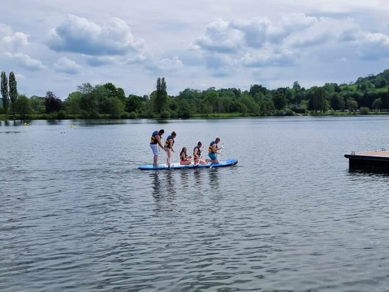 Groupe de personnes debout sur un grand paddle board bleu sur un lac sous un ciel nuageux.