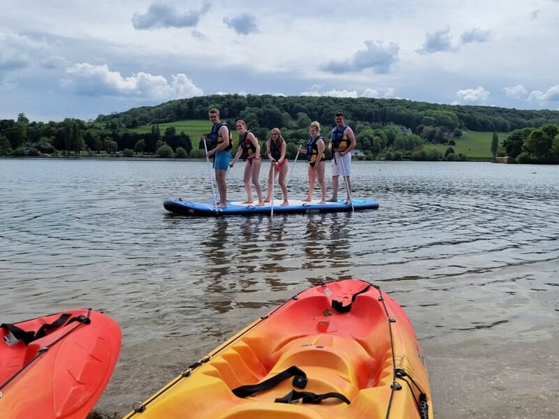 Cinq personnes debout sur un paddle board bleu sur un lac, avec kayaks orange au premier plan.