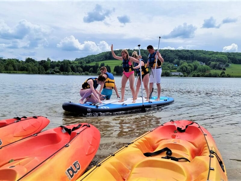 Groupe de personnes s'amusant sur un paddle board bleu au bord d'un lac avec kayaks orange.