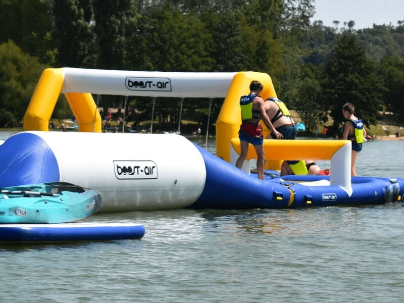 Personnes en gilets de sauvetage sur un parc aquatique flottant, évoluant autour d'obstacles gonflables, dans un cadre naturel avec des arbres et une colline en arrière-plan