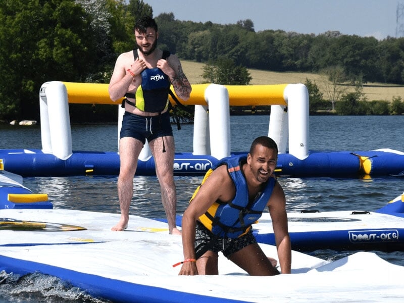 Deux hommes se tenant debout sur un module gonflable, l'un en équilibre instable, l'autre souriant, partageant un moment de joie et d'activité physique sur l'eau