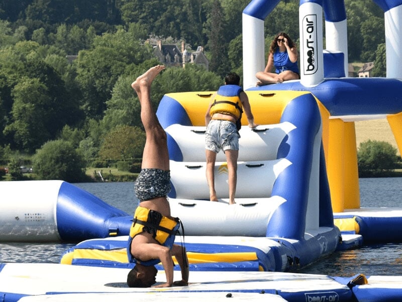 Activité ludique sur une structure aquatique, avec des participants en gilets de sauvetage, certains marchant, d'autres tombant à l'eau, dans un environnement estival et dynamique