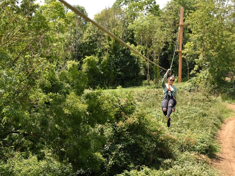 Une personne faisant de la tyrolienne à travers une forêt verdoyante. Elle porte des lunettes de soleil et est attachée à un harnais, profitant de l'aventure en pleine nature