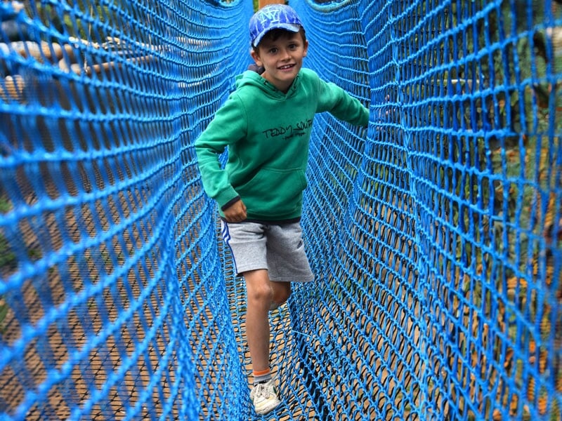 Un garçon souriant, vêtu d'un sweat à capuche vert et d'un short gris, traversant un pont suspendu en filet bleu dans un parcours aventure en plein air. Il porte une casquette bleue et semble s'amuser