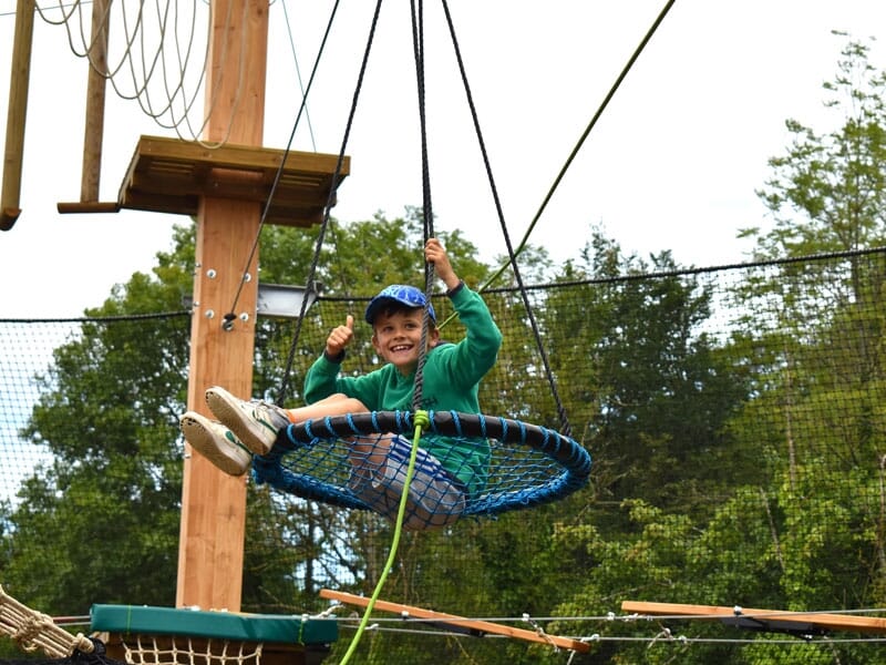 Un garçon assis dans une grande balançoire en filet bleu, suspendue dans un parcours d'accrobranche. Il porte un sweat à capuche vert et une casquette bleue, et affiche un grand sourire