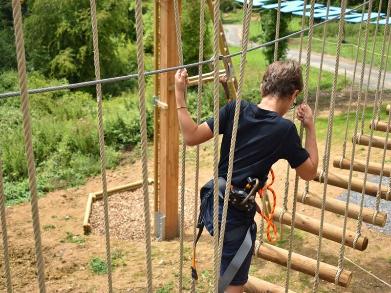 Une personne portant un t-shirt noir et un harnais de sécurité traversant un parcours d'accrobranche. Elle est concentrée sur son équilibre en tenant les cordes de sécurité. Le sol en dessous est cons