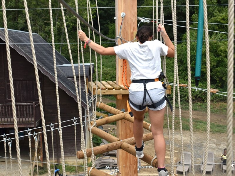 Une personne traversant un pont de corde dans un parcours d'accrobranche, portant un t-shirt noir et un harnais de sécurité. Le terrain en dessous est légèrement en pente et verdoyant