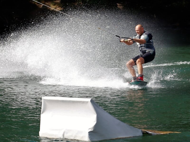 Homme en wakeboard tiré par un câble, éclaboussant l'eau en effectuant un saut sur un obstacle.