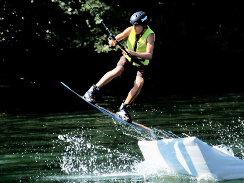 Enfant en wakeboard portant un casque et un gilet de sauvetage, sautant sur un obstacle sur l'eau.